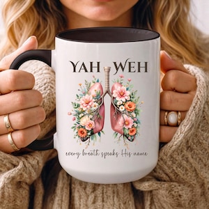 May include: White ceramic mug with a black handle and rim, featuring the words "YAH WEH" above a watercolor illustration of lungs adorned with flowers. The phrase "every breath speaks His name" is written below the lungs.