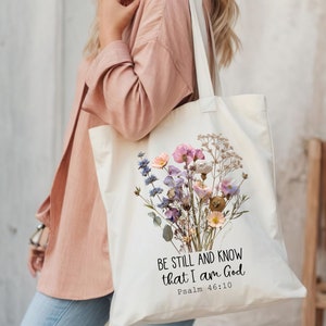 May include: A white canvas tote bag with a floral design and the text "Be still and know that I am God, Psalm 46:10".