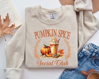 May include: Beige sweatshirt with the text "PUMPKIN SPICE Social Club" in orange. The graphic includes a pumpkin, a pumpkin spice latte, and fall foliage. The sweatshirt is folded on a white surface.