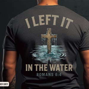 Op de afbeelding: Donkergrijs t-shirt met de tekst "I LEFT IT IN THE WATER" en "ROMANS 6:4". Het shirt heeft een houten kruis in water.