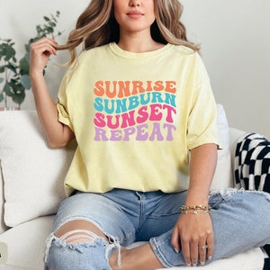 Op de afbeelding: Lichtgele t-shirt met de tekst "Sunrise Sunburn Sunset Repeat" in kleurrijke retroletters. Het shirt wordt gedragen met een blauwe jeans. De algehele uitstraling is casual en zomers.