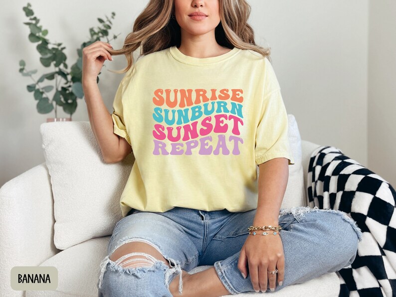 Op de afbeelding: Lichtgele t-shirt met de tekst "Sunrise Sunburn Sunset Repeat" in kleurrijke retroletters. Het shirt wordt gedragen met een blauwe jeans. De algehele uitstraling is casual en zomers.