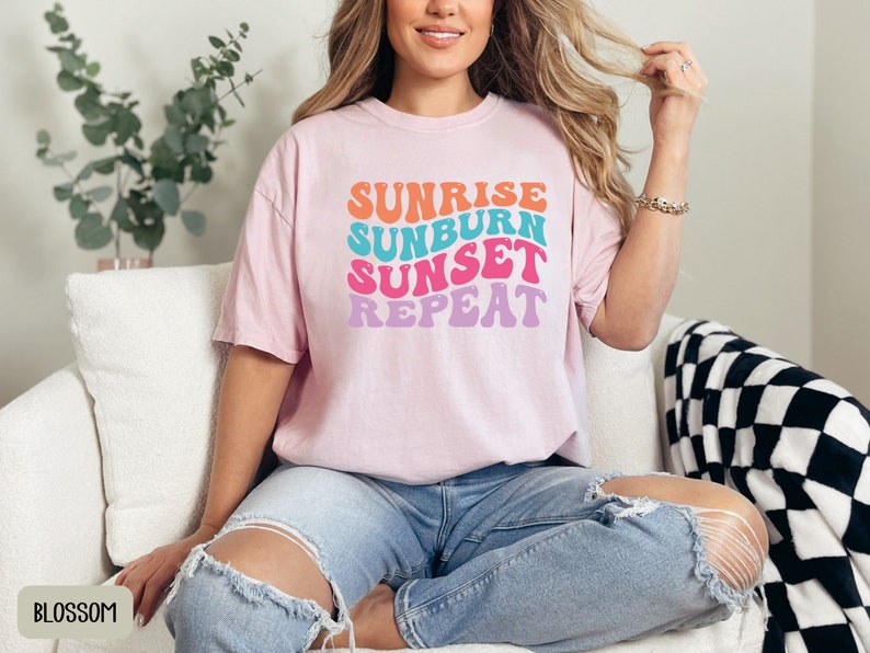 Op de afbeelding: Een lichtroze t-shirt met de tekst "Sunrise Sunburn Sunset Repeat" in een retro lettertype. De tekst is in oranje, turquoise, roze en paars. Het shirt wordt gecombineerd met een blauwe jeans.