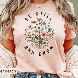 May include: A heather peach t-shirt with a floral design and the text "Be Still and Know".