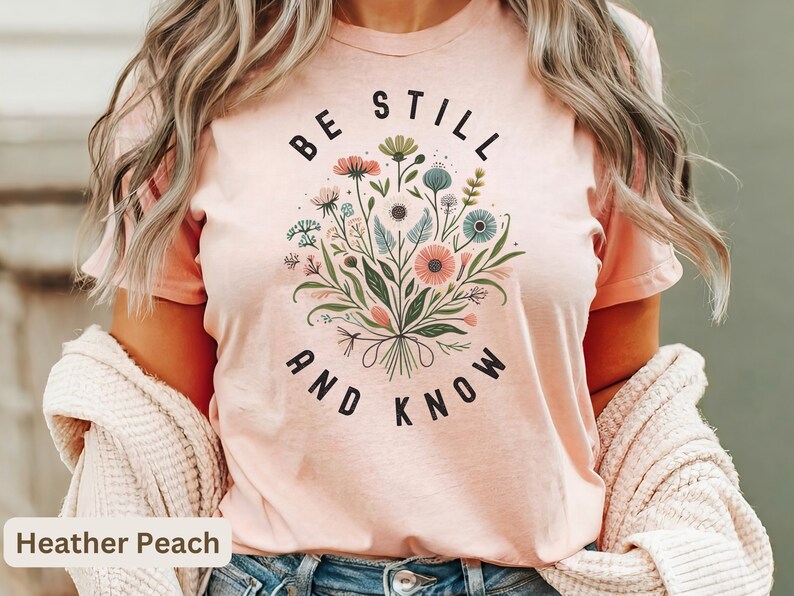 May include: A heather peach t-shirt with a floral design and the text "Be Still and Know".
