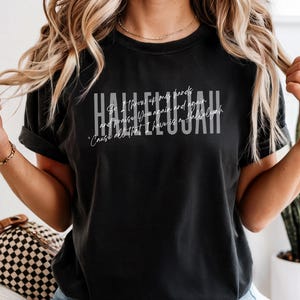May include: A black short-sleeve t-shirt featuring the word HALLELUJAH in large white block letters. Overlaid in a thin white script font is the text: So I throw up my hands and praise You again and again. 'Cause all that I have is a Hallelujah.