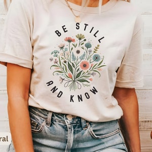 May include: A white t-shirt with a floral design and the text "Be Still and Know" printed on the front. The shirt is being worn by a person with a necklace.