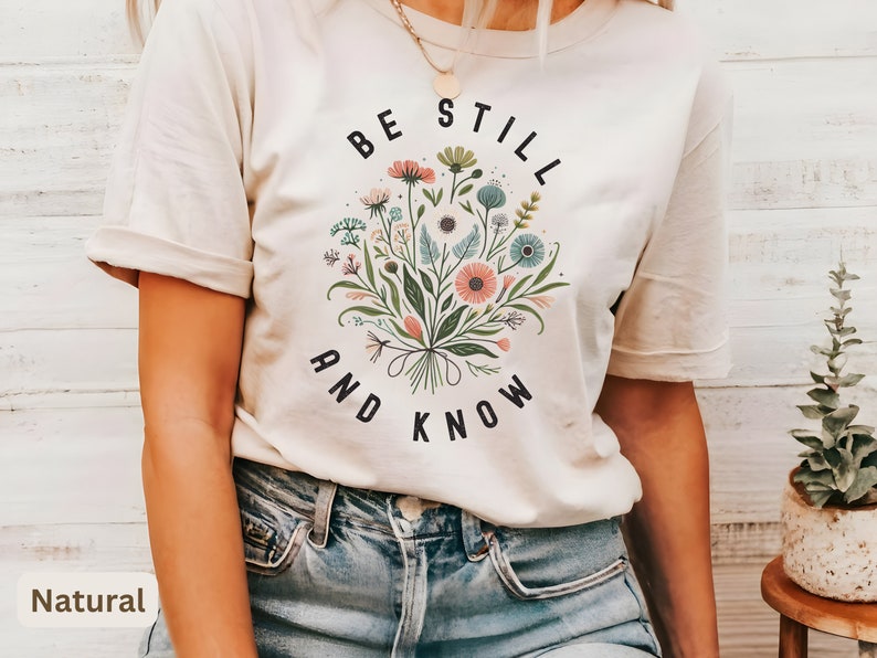May include: A white t-shirt with a floral design and the text "Be Still and Know" printed on the front. The shirt is being worn by a person with a necklace.