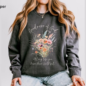 May include: Dark gray sweatshirt with a floral bouquet design in shades of orange, pink, and blue. The text "Goodness of God" and "All my life you have been faithful" are written in a script font. The sweatshirt is a comfortable, casual style.