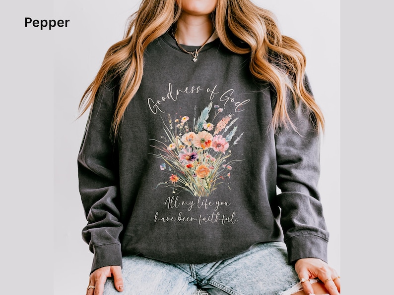 May include: Dark gray sweatshirt with a floral bouquet design in shades of orange, pink, and blue. The text "Goodness of God" and "All my life you have been faithful" are written in a script font. The sweatshirt is a comfortable, casual style.