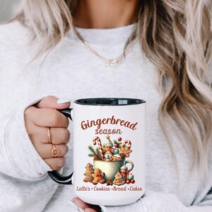 May include: White ceramic mug with a black interior and handle, featuring a "Gingerbread Season" design. The design includes gingerbread cookies, candy canes, and other festive elements. The mug also lists "Latte's &bull; Cookies &bull; Bread &bull; Cakes".