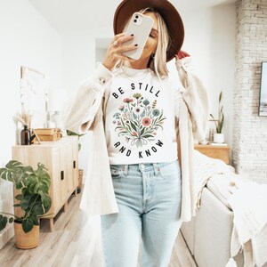 May include: White t-shirt with a floral design and the text "Be Still and Know" printed on the front.