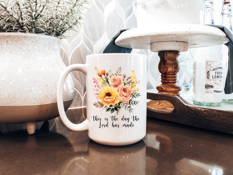 Puede incluir: Taza de cer&aacute;mica blanca con un dise&ntilde;o floral y el texto "This is the day the Lord has made".