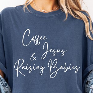 以下が含まれることがあります： デニムブルーのTシャツ。白い筆記体で「Coffee & Jesus Raising Babies」と書かれています。 ゆったりとしたフィット感で、袖はロールアップされています。 左下に「DENIM」の文字が小さなボックスで印刷されています。