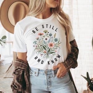 May include: White t-shirt with a floral bouquet graphic and the text "Be Still and Know" in black.