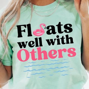 May include: A light green t-shirt with the text "Floats well with Others" in black and pink lettering. A pink flamingo pool float is above the word "Floats".