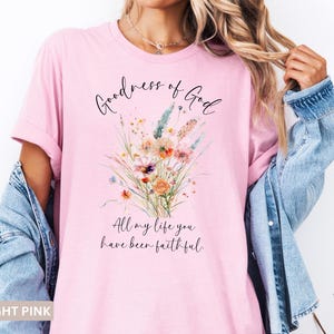 May include: Light pink t-shirt with the text "Goodness of God" and "All my life you have been faithful." A watercolor floral design in orange, blue, and yellow is printed on the front. The shirt is worn with a denim jacket.
