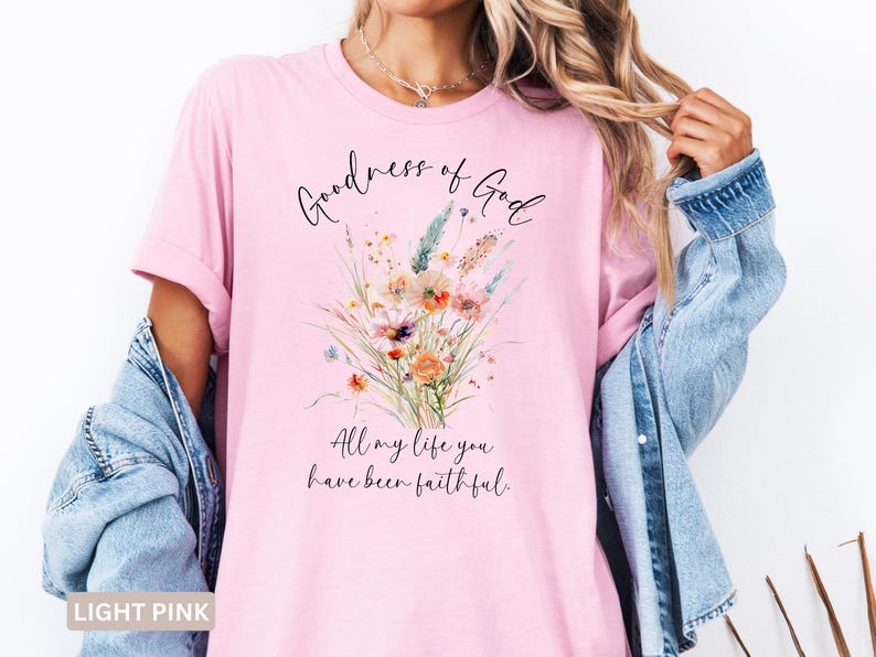 May include: Light pink t-shirt with the text "Goodness of God" and "All my life you have been faithful." A watercolor floral design in orange, blue, and yellow is printed on the front. The shirt is worn with a denim jacket.