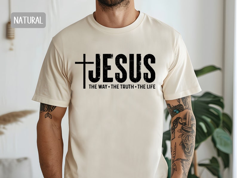 May include: A beige t-shirt with a black graphic that says "JESUS THE WAY - THE TRUTH - THE LIFE" with a cross before the word "JESUS".