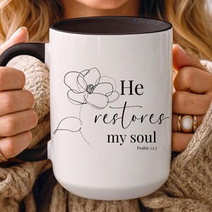 May include: White ceramic mug with a black handle and interior. The mug features a black line drawing of a flower and the text "He restores my soul" with the Bible verse Psalm 23:3.