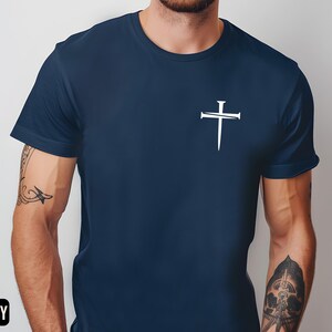 May include: A navy blue t-shirt with a white cross design featuring nails. The cross is centered on the chest of the shirt.