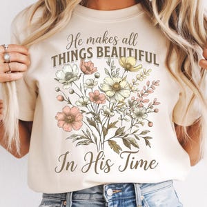 May include: Cream-colored t-shirt featuring a floral bouquet design with pink, white, and yellow flowers. The text "He makes all THINGS BEAUTIFUL In His Time" is printed above the flowers in a script font.