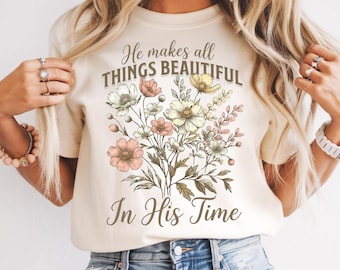 May include: Cream-colored t-shirt featuring a floral bouquet design with pink, white, and yellow flowers. The text "He makes all THINGS BEAUTIFUL In His Time" is printed above the flowers in a script font.
