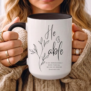 May include: White ceramic mug with a black handle and interior. The mug features the script "He is able" with a floral design and the text "To do immeasurably more than all we ask or imagine Ephesians 3:20."