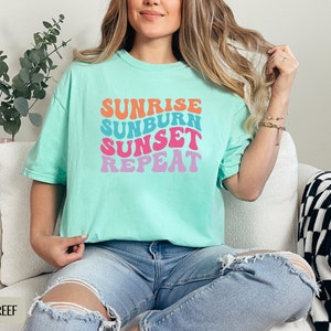 Op de afbeelding: Lichtgroen t-shirt met de tekst "SUNRISE SUNBURN SUNSET REPEAT" in retro-stijl, kleurrijke letters. De tekst is in oranje, blauw, roze en paars. Het shirt wordt gedragen met een blauwe jeans. Casual stijl.