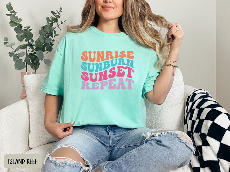 Op de afbeelding: Lichtgroen t-shirt met de tekst "SUNRISE SUNBURN SUNSET REPEAT" in retro-stijl, kleurrijke letters. De tekst is in oranje, blauw, roze en paars. Het shirt wordt gedragen met een blauwe jeans. Casual stijl.