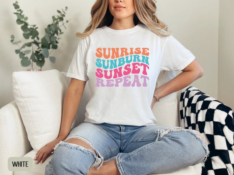 Op de afbeelding: Wit T-shirt met de tekst "Sunrise Sunburn Sunset Repeat" in oranje, turquoise, roze en paars. Het shirt wordt gedragen door een persoon die een blauwe spijkerbroek draagt. De persoon zit op een witte bank. Een zwart-wit geblokte deken ligt op de bank.