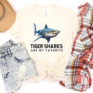 May include: A beige t-shirt with a graphic of a tiger shark and the text "Tiger Sharks Are My Favorite". The shirt is displayed on a white background with a pair of denim shorts, a straw hat, and a plaid shirt.