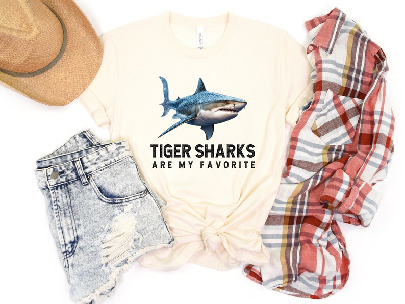 May include: A beige t-shirt with a graphic of a tiger shark and the text "Tiger Sharks Are My Favorite". The shirt is displayed on a white background with a pair of denim shorts, a straw hat, and a plaid shirt.