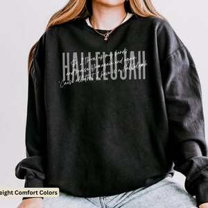 May include: A black long-sleeve crewneck sweatshirt featuring the word HALLELUJAH in large white block letters. Overlaid in a smaller white script font is the text: So I throw up my hands and praise You again and again 'Cause all that I have is a Hallelujah.