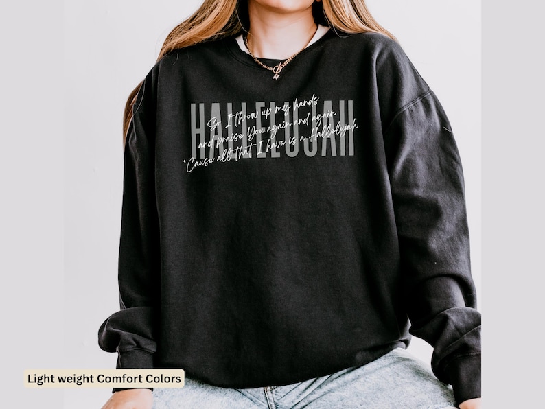 May include: A black long-sleeve crewneck sweatshirt featuring the word HALLELUJAH in large white block letters. Overlaid in a smaller white script font is the text: So I throw up my hands and praise You again and again 'Cause all that I have is a Hallelujah.