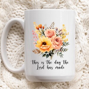 Puede incluir: Taza de cer&aacute;mica blanca con un dise&ntilde;o floral con flores amarillas, naranjas y rosas. El texto "This is the day the Lord has made" est&aacute; impreso en la taza.
