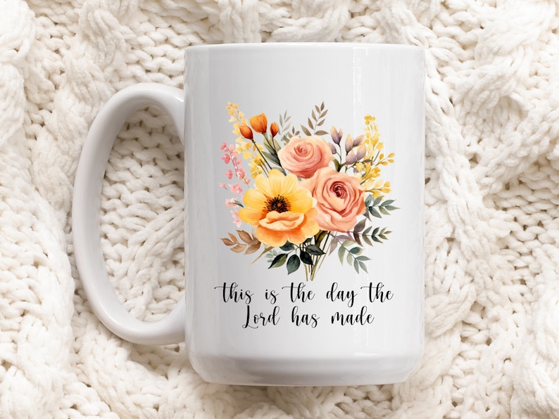 Puede incluir: Taza de cer&aacute;mica blanca con un dise&ntilde;o floral con flores amarillas, naranjas y rosas. El texto "This is the day the Lord has made" est&aacute; impreso en la taza.