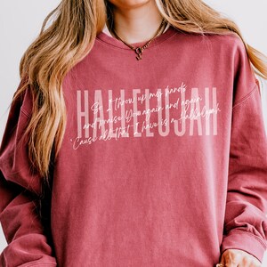 May include: A crimson crewneck sweatshirt featuring the word HALLELUJAH in large white block letters. Overlaid is the white cursive text: So I throw up my hands and praise You again and again 'Cause all that I have is a Hallelujah.