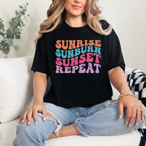 Op de afbeelding: Zwart T-shirt met de tekst "SUNRISE SUNBURN SUNSET REPEAT" in oranje, blauw, roze en paars. Het shirt wordt gedragen met een blauwe spijkerbroek. Het model zit op een witte bank.