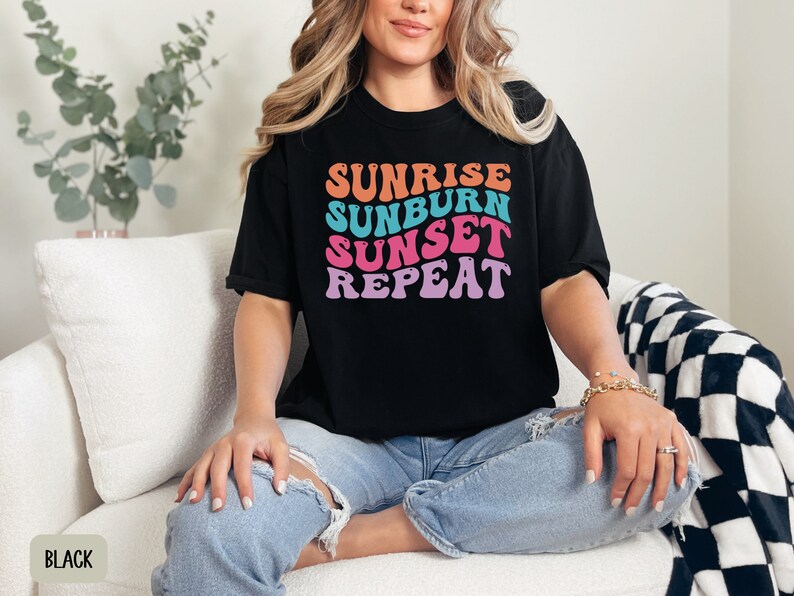 Op de afbeelding: Zwart T-shirt met de tekst "SUNRISE SUNBURN SUNSET REPEAT" in oranje, blauw, roze en paars. Het shirt wordt gedragen met een blauwe spijkerbroek. Het model zit op een witte bank.