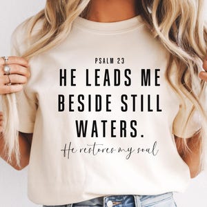 May include: Ivory-colored t-shirt with the text "Psalm 23 He leads me beside still waters. He restores my soul" in black. The shirt is a crew neck style.
