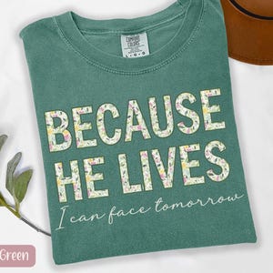May include: Light green t-shirt with the text "BECAUSE HE LIVES" in large, floral-patterned letters. Below, the shirt reads "I can face tomorrow" in a script font. A brown hat is visible in the background.