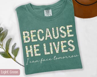 May include: Light green t-shirt with the text "BECAUSE HE LIVES" in large, floral-patterned letters. Below, the shirt reads "I can face tomorrow" in a script font. A brown hat is visible in the background.