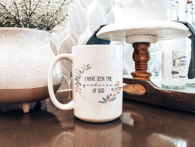 May include: White ceramic coffee mug with a gold rim and floral design. The mug reads "I have seen the goodness of God".