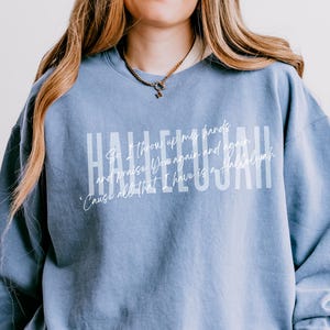 May include: A blue jean colored crewneck sweatshirt featuring the word HALLELUJAH in large white block letters. Overlaid in a smaller white script font is the text: So I throw up my hands and praise You again and again, 'Cause all that I have is a Hallelujah.