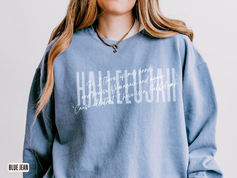 May include: A blue jean colored crewneck sweatshirt featuring the word HALLELUJAH in large white block letters. Overlaid in a smaller white script font is the text: So I throw up my hands and praise You again and again, 'Cause all that I have is a Hallelujah.