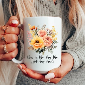 Puede incluir: Taza de cer&aacute;mica blanca con un dise&ntilde;o floral con flores amarillas, rosas y verdes. La taza tiene el texto "This is the day the Lord has made" impreso en la parte inferior.