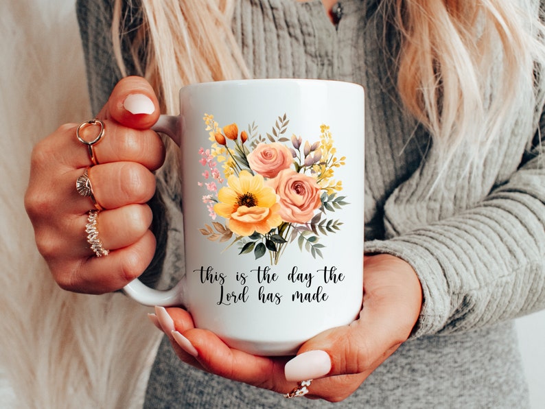 Puede incluir: Taza de cer&aacute;mica blanca con un dise&ntilde;o floral con flores amarillas, rosas y verdes. La taza tiene el texto "This is the day the Lord has made" impreso en la parte inferior.