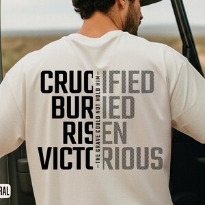 Op de afbeelding: Crèmekleurig T-shirt met de woorden "CRUCIFIED, BURIED, RISEN, VICTORIOUS" in een vet, zwart lettertype. De woorden zijn verticaal gestapeld. De zin "THE GRAVE COULD NOT HOLD HIM" is verticaal gedrukt.