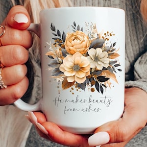 May include: White ceramic mug with a floral design in shades of yellow, brown, and gray. The mug has a handle and the text "He makes beauty from ashes" printed on the front.